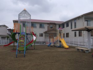 2階建ての建物が建つ隣に広がる、カラフルなすべり台のような遊具が設置された、土の地面の公園の写真