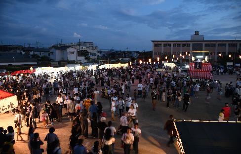 沢山の人々が、複数の夜店やひとつの櫓の周りに集まり、賑わいを見せる盆踊りの夜の写真