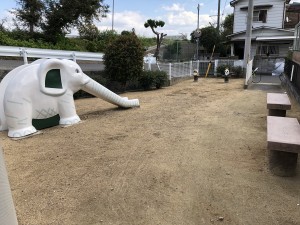 奥に民家が見える、地面が土の広場に、白い象の形をしたすべり台とベンチが2基並んでいる写真