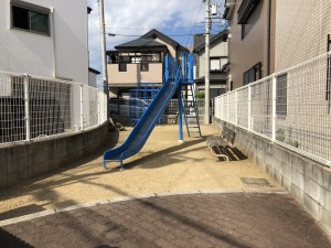 建物に挟まれた公園の広場に設置された青色のすべり台の写真