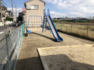 すべり台、ブランコ、ベンチが設置されている道路沿いの広場の写真