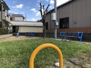 建物に囲まれ、土の地面と緑色の草が広がる広場に、U字型の黄色い鉄のオブジェと、青色のベンチが2基、シーソーが1台設置されている写真