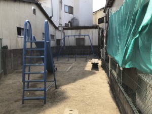 周りを建物に囲まれた敷地にブランコと滑り台がある広場の写真