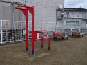 白いフェンスの前に茶色のベンチが2つ並び手前に赤色の健康遊具が設置されている写真