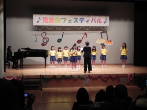 児童館フェスティバルの舞台上で、ピアノの横で黄色いシャツを着た合唱教室のこどもたちが先生の指揮に合わせて合唱している様子の写真