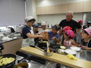 調理場でエプロンと三角巾をつけてジャガイモ料理に挑戦する子どもたちの写真