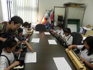 大正琴の楽譜を見ながら練習しているこどもと後ろから先生が楽譜を指さしながら教えている様子の写真
