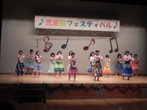 児童館フェスティバルの舞台上で、ひらひらで色とりどりのスカートを履いた子どもたちが両手を胸の前でバツ印に交差している写真