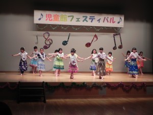 児童館フェスティバルの舞台上で、ひらひらで色とりどりのスカートを履いた子どもたちが両手を広げるようなポーズでフラダンスを披露している写真