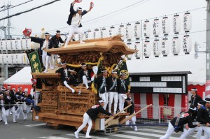 だんじりの一番上でうちわを持って両手を挙げている人やだんじり曳行する法被姿の人たちの写真