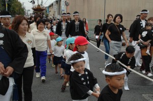 だんじりを引っ張る体操服や法被を着た子供たちとそれを近くで見守る大人たちの画像