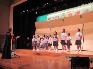 第27回町民音楽祭と書かれた横断幕がある舞台上で指揮者の指揮に合わせて歌う児童合唱教室バンビーナの子どもたちの写真