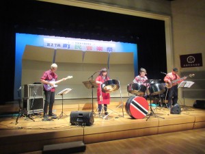 第27回町民音楽祭と書かれた横断幕がある舞台上で演奏する釣 千賀子さんとSteeling Beatのメンバーたちの写真
