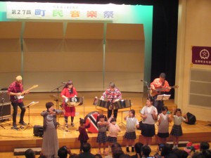 第27回町民音楽祭と書かれた横断幕がある舞台上で演奏する釣 千賀子さんとSteeling Beatのメンバーたちと舞台前で一緒に演奏に花を添える今回の参加者たちの写真