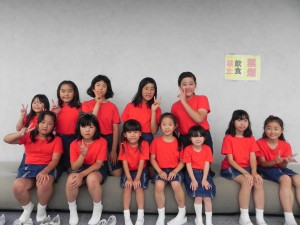 赤いチームカラーのシャツを着た12人の子どもたちが2列に並んで座り、カメラに向かってピースなどのポーズをとっている「こども合唱」教室の写真