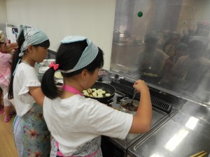 輪切りのジャガイモをフライパンで炒める少女とその横で調味料を合わせたおだしを小さな鍋で煮る少女の写真