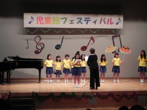 児童館フェスティバルの舞台上で、ピアノの横で黄色いシャツを着た合唱教室のこどもたちの発表風景の写真