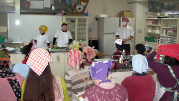 調理実習室でエプロンとスカーフ着用の人たちが講師から話を聞いている写真